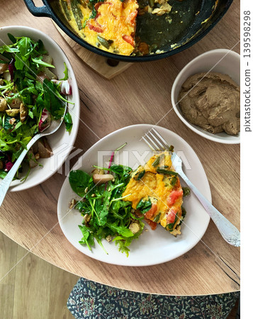 Baked vegetables with eggs sit on a plate next to a salad made from arugula and Brussels sprouts. A bowl of liver pate completes the meal with healthy choices Baked vegetables with eggs sit on a plate next to a salad made from arugula and Brussels sprouts. A bowl of liver pate completes the meal with healthy choices 139598298