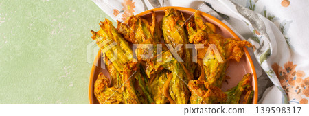 Golden-brown deep-fried zucchini flowers are arranged on a vibrant plate. The dish is set on a floral-patterned napkin, enhancing its appetizing appearance, banner Golden-brown deep-fried zucchini flowers are arranged on a vibrant plate. The dish is set on a floral-patterned napkin, enhancing its appetizing appearance, banner 139598317