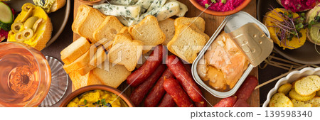 A vibrant charcuterie board showcases a variety of cheese, sausages, hummus, nuts, and sandwiches. Friends gather at a sunny outdoor picnic, enjoying delicious food together, banner A vibrant charcuterie board showcases a variety of cheese, sausages, hummus, nuts, and sandwiches. Friends gather at a sunny outdoor picnic, enjoying delicious food together, banner 139598340