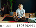 Middle aged woman using phone for setting mobile app for meditation at home, sitting on yoga mat in modern dark Scandi interior home with green plants. Meditating, relaxing practice at home. 139598441