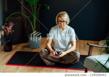 Middle aged woman using phone for setting mobile app for meditation at home, sitting on yoga mat in modern dark Scandi interior home with green plants. Meditating, relaxing practice at home. Middle aged woman using phone for setting mobile app for meditation at home, sitting on yoga mat in modern dark Scandi interior home with green plants. Meditating, relaxing practice at home. 139598441