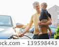 Young man holding baby and install cable plug to his electric car with home charging station. Residential EV Charging Station. Sustainable Power of a Wall-Mounted Charger. Future of Clean Transport. 139598443