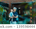 Young father holding toddler son and watering plants in domestic garden backyard at home. Child watering plants from a hose. Routine household chores with baby. Time together. 139598444