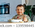 Happy smiling cute toddler dirty baby eating himself from plate in the kitchen. Funny boy in feeding bib self-feeding solid food fine motor development. Everyday cooking and Baby's feeding process. 139598445