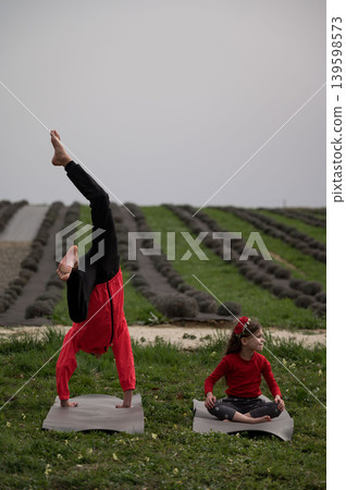 Two children practicing yoga in a serene field surrounded by neatly arranged rows of plants while one performs a handstand and the other sits calmly in meditation during the golden hour Two children practicing yoga in a serene field surrounded by neatly arranged rows of plants while one performs a handstand and the other sits calmly in meditation during the golden hour 139598573
