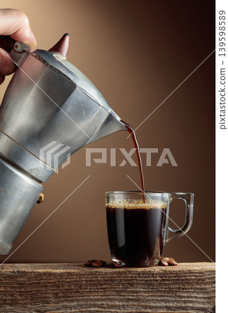 Black coffee is poured from a classic Italian aluminium moka pot into a glass coffee cup. Black coffee is poured from a classic Italian aluminium moka pot into a glass coffee cup. 139598589