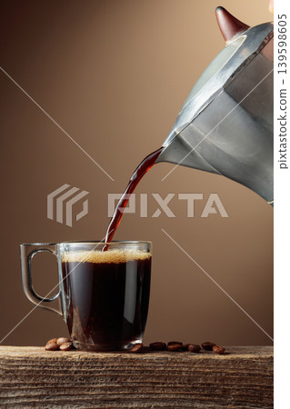 Black coffee is poured from a classic Italian aluminium moka pot into a glass coffee cup. Black coffee is poured from a classic Italian aluminium moka pot into a glass coffee cup. 139598605