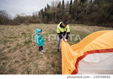Camping adventure unfolds as a father teaches his daughter the art of tent setup amidst a serene landscape under overcast skies in early spring 139598660