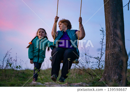 Grandmother and granddaughter share a joyful swing ride at sunset. Precious moments of intergenerational connection. Grandmother and granddaughter share a joyful swing ride at sunset. Precious moments of intergenerational connection. 139598687