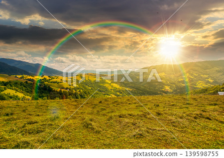 sunset over mountain landscape with grassy field. trees on the rolling hills and rural valley beneath a cloudy sky with rainbow in the background. view of carpathian mountains in summer 139598755