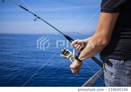 Close-up of man's hand holding fishing rod in the sea, man fishing. Fishing rod with a spinning reel in the hands of a fisherman. Close-up of man's hand holding fishing rod in the sea, man fishing. Fishing rod with a spinning reel in the hands of a fisherman. 139600106