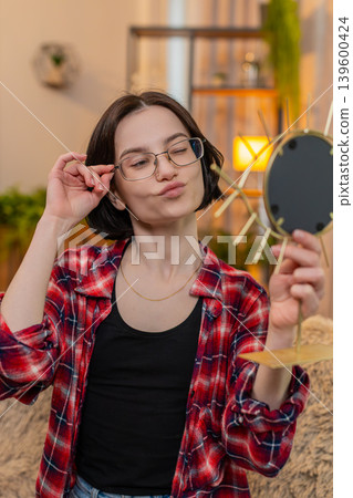 Young woman trying on new glasses at mirror, smiling while checking reflection with satisfaction Young woman trying on new glasses at mirror, smiling while checking reflection with satisfaction 139600424