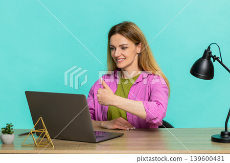 Adult woman sitting at table showing thumbs up to laptop webcam giving positive feedback with smile Adult woman sitting at table showing thumbs up to laptop webcam giving positive feedback with smile 139600481