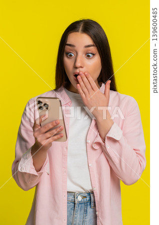 Woman looking surprised at smartphone suddenly covering mouth shocked unexpected surprise reaction 139600485
