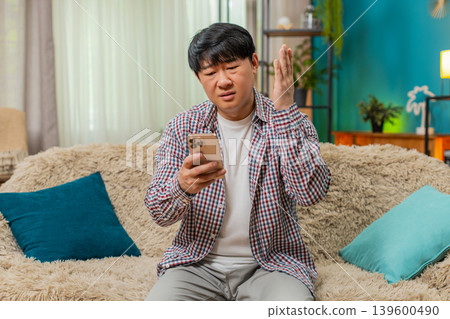 Asian man frowning at smartphone, disappointed expression, frustrated after reading bad news at home 139600490