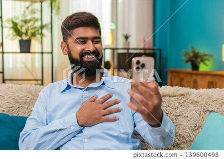Indian man smiles gently hand on heart viewing touching nostalgic photo on smartphone memory indoors Indian man smiles gently hand on heart viewing touching nostalgic photo on smartphone memory indoors 139600498