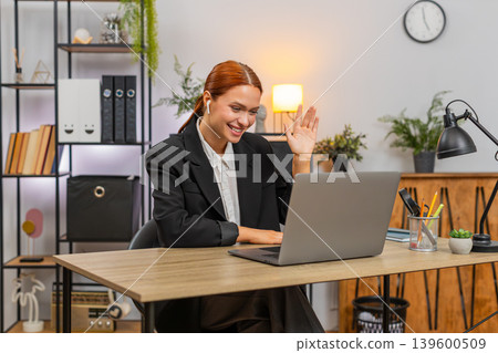 Young businesswoman joins video call on laptop wears earphones waves hi and gestures energetically Young businesswoman joins video call on laptop wears earphones waves hi and gestures energetically 139600509