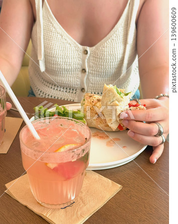 Hands of woman in summer top eating wrap lunch at cafe table with lemonade, salad, plate. Concept of lunch break, casual dining, candid lifestyle, refreshment, snack Hands of woman in summer top eating wrap lunch at cafe table with lemonade, salad, plate. Concept of lunch break, casual dining, candid lifestyle, refreshment, snack 139600609