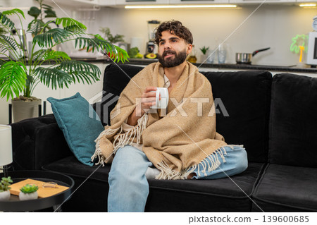 Indian man holding warm tea cup wrapped in blanket slowly sipping drink relaxing with gentle smile 139600685