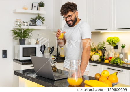 Arabian young adult man drinking orange juice reading news on laptop in home kitchen morning routine 139600699