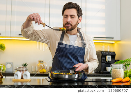 Middle-aged man tasting vegetables on skillet pan at home kitchen cooking healthy diet meal on stove Middle-aged man tasting vegetables on skillet pan at home kitchen cooking healthy diet meal on stove 139600707