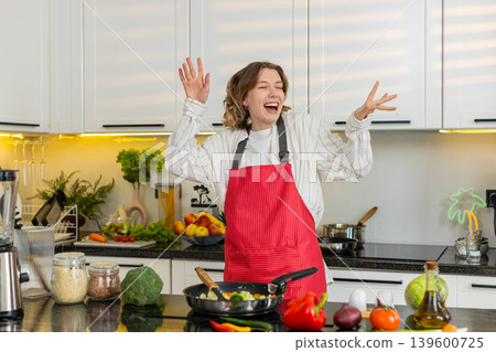 Young woman dancing and singing while cooking healthy vegetables in kitchen for diet lunch at home Young woman dancing and singing while cooking healthy vegetables in kitchen for diet lunch at home 139600725