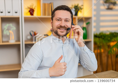 Young man on home table working in headset with laptop and smiling during virtual customer support Young man on home table working in headset with laptop and smiling during virtual customer support 139600734