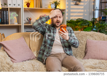 Young man on home sofa shocked by order mistake on smartphone and touching head in worried reaction Young man on home sofa shocked by order mistake on smartphone and touching head in worried reaction 139600735