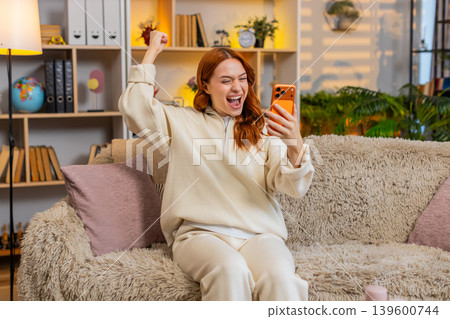 Young woman on home sofa reading smartphone message and celebrating online win with joyful triumph Young woman on home sofa reading smartphone message and celebrating online win with joyful triumph 139600744