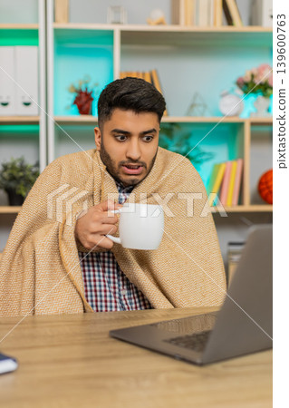 Indian man on home table warming with hot drink under blanket while working on laptop in cold room 139600763