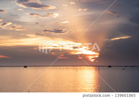 Gentle sunset light spreads across a wide lake, illuminating soft clouds and creating a warm golden reflection on calm water with distant structures along the horizon. 139601865