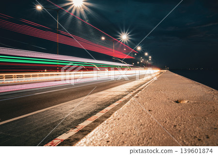 Long exposure night scene capturing vibrant light trails from moving vehicles across a bridge, illuminated by street lamps, creating dynamic motion lines and a sense of speed and urban energy. 139601874
