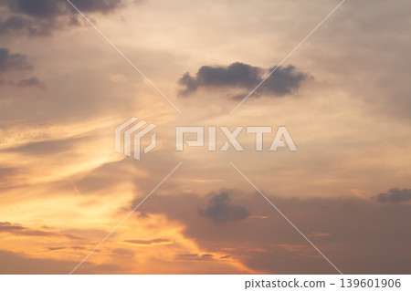 Warm sunset colors blending with drifting dark clouds and glowing light patterns across the evening sky, forming a peaceful atmospheric background. 139601906