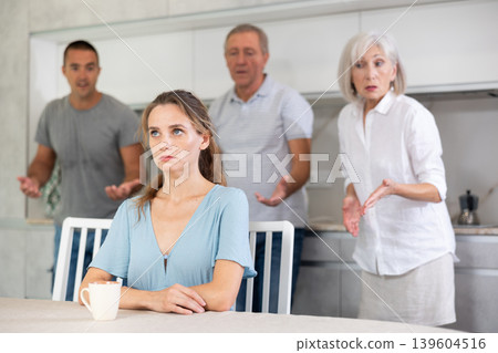 Domestic quarrel - offended young woman sits at table while parents and young husband yell at her Domestic quarrel - offended young woman sits at table while parents and young husband yell at her 139604516