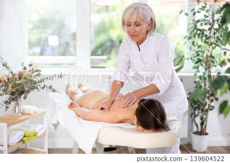 Skilled aged masseuse doing back pain relief massage to young woman Skilled aged masseuse doing back pain relief massage to young woman 139604522