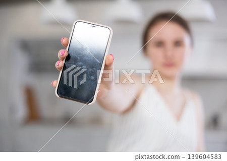 In home kitchen with white interior, smiling European girl in dress shows blank smartphone screen In home kitchen with white interior, smiling European girl in dress shows blank smartphone screen 139604583