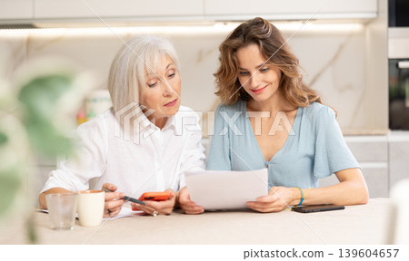 Positive woman and her senior mother sitting at table in kitchen, doing family financial paperwork 139604657