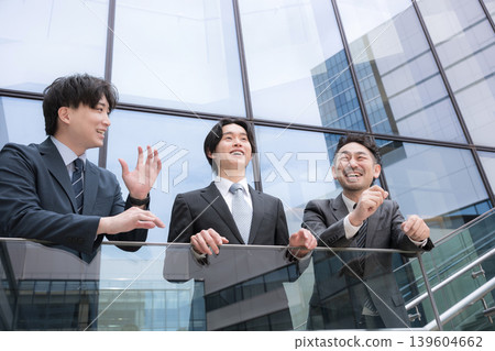 The image of a three-person team, with a positive and forward-thinking boss watching over a younger subordinate, is set in front of a building outdoors. 139604662