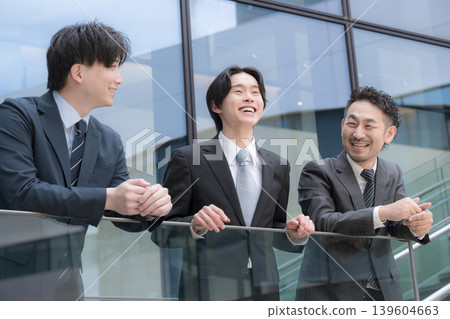The image of a three-person team, with a positive and forward-thinking boss watching over a younger subordinate, is set in front of a building outdoors. 139604663
