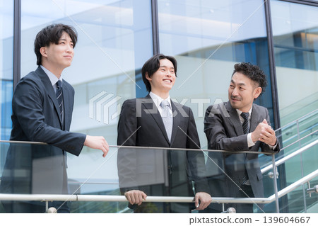 The image of a three-person team, with a positive and forward-thinking boss watching over a younger subordinate, is set in front of a building outdoors. The image of a three-person team, with a positive and forward-thinking boss watching over a younger subordinate, is set in front of a building outdoors. 139604667