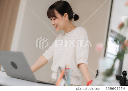 Happy smiling asian woman using modern technology computer working standing at bright desk inside home office room Happy smiling asian woman using modern technology computer working standing at bright desk inside home office room 139605254
