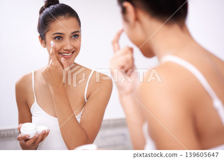 Woman, skincare and moisturizing in bathroom mirror, lotion and face cream on skin for hydration. Happy female person, reflection and cosmetics or dermatology, self care and apply sunblock at home 139605647
