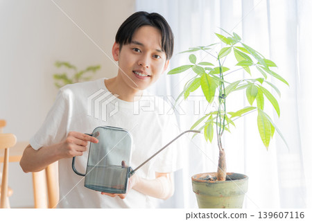 A young man waters a houseplant with a watering can. A young man waters a houseplant with a watering can. 139607116