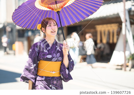 A woman in a yukata (summer kimono) and carrying a traditional Japanese umbrella enjoys sightseeing in the historic town of Kawagoe, a summer travel destination, strolling through the historic storehouse-lined streets. A woman in a yukata (summer kimono) and carrying a traditional Japanese umbrella enjoys sightseeing in the historic town of Kawagoe, a summer travel destination, strolling through the historic storehouse-lined streets. 139607726