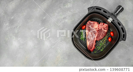 Raw porterhouse t bone steak in air fryer basket with herbs on gray background Raw porterhouse t bone steak in air fryer basket with herbs on gray background 139608771