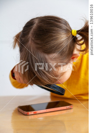 A little girl examines a mobile phone with a magnifying glass, introducing concepts such as early exposure to mobile phones or internet safety. 139610016