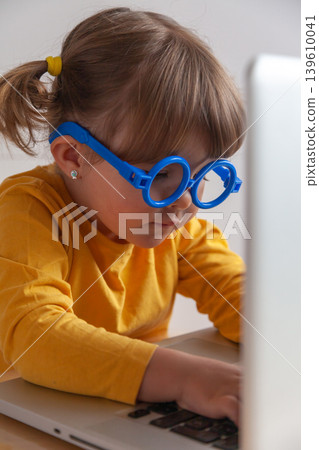 Little girl with blue glasses staring at a computer screen introducing concepts such as effects of screen time on an eye health. 139610041