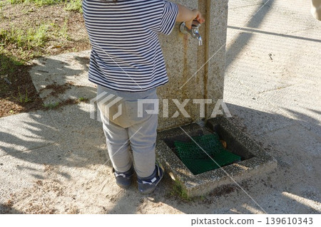 A child's back as they turn on the tap at a drinking fountain in a park. A child's back as they turn on the tap at a drinking fountain in a park. 139610343