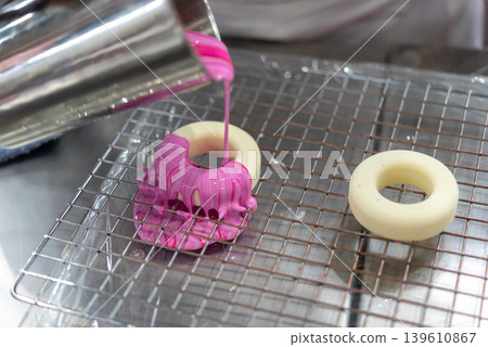 Fresh donut being coated with pink glaze on cooling rack during bakery preparation process with sweet dessert decoration detail 139610867