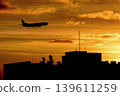 Airplane silhouette landing above city skyline at sunset with dramatic orange sky and rooftop antennas creating urban travel atmosphere. 139611259